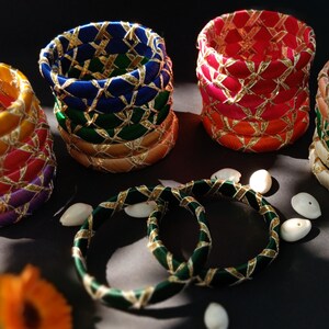May include: A collection of circular bangles wrapped in colorful silk thread, including yellow, blue, red, purple, green, orange, and beige. Each bangle features a decorative crisscross pattern of metallic gold thread wrapped around the exterior.