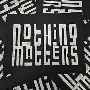 Op de afbeelding: Zwarte rechthoekige kaarten met de woorden "NOTHING MATTERS" in grote, verweerde witte blokletters. De kaarten zijn verspreid, met de zin herhaald op sommige. Het algehele effect is vet en grafisch.
