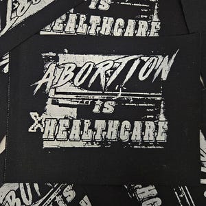 May include: Black rectangular patch with the words "ABORTION IS HEALTHCARE" in distressed white lettering. The text is arranged in two lines, with "ABORTION" above "IS HEALTHCARE". Several patches are visible.
