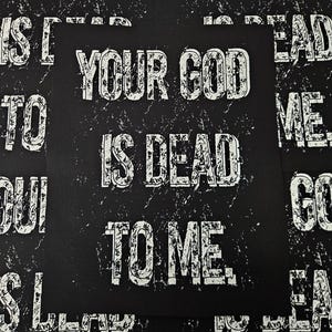 Op de afbeelding: Een zwarte patch met witte tekst die "Your God Is Dead To Me" luidt. De tekst is in een vetgedrukt, bloklettertype en heeft een verweerde, versleten uitstraling.