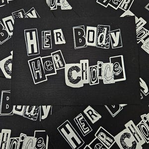 May include: Black cards with the text "HER BODY HER CHOICE" in a white, distressed font. The cards are scattered, with the text visible on several. The overall theme is related to women's rights and reproductive freedom.