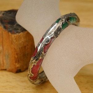 May include: A silver-toned metal bangle bracelet featuring alternating inlaid sections of red and green enamel. The metal surface has a textured, hammered appearance with raised edges surrounding each colored enamel segment.