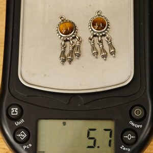 May include: A pair of silver earrings with brown tiger's eye stones and dangling tassels. The earrings are on a digital scale that reads 5.7 grams.