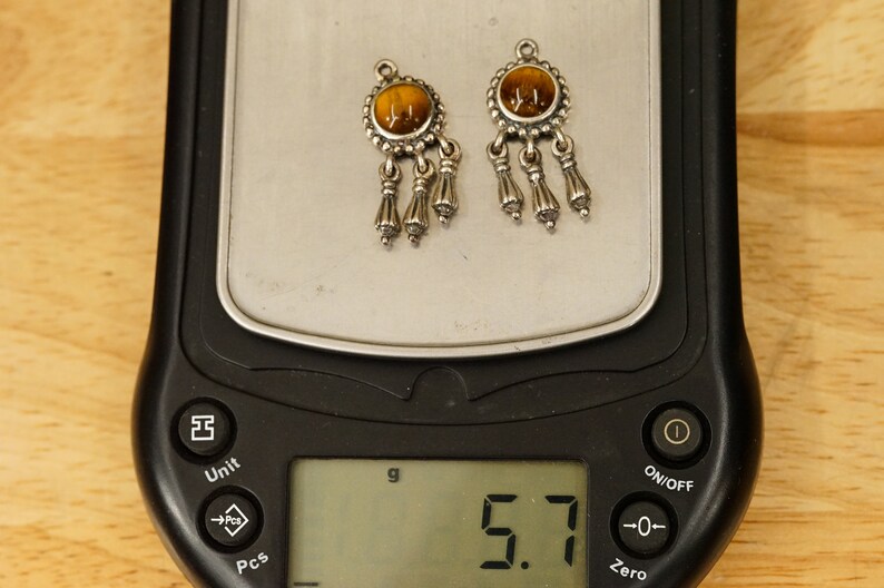May include: A pair of silver earrings with brown tiger's eye stones and dangling tassels. The earrings are on a digital scale that reads 5.7 grams.