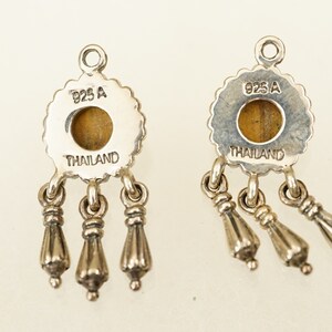 May include: A pair of silver earrings with a tiger's eye stone and three dangling charms. The back of the earrings are stamped with "925 A THAILAND".