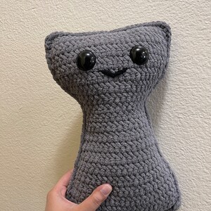 May include: A gray crocheted plush toy shaped like a cat with black button eyes and a stitched smile.