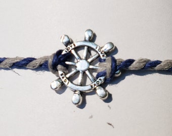 May include: A silver-colored ship's wheel charm with the word "love" inscribed on each spoke, tied to a braided rope bracelet with blue and gray strands.