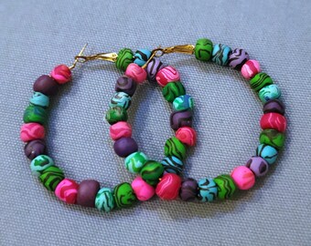 May include: A pair of hoop earrings made of colorful beads. The beads are green, pink, purple, and blue with a swirl pattern. The earrings are connected by a gold metal hoop.