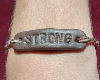 May include: A brown leather bracelet with the word "STRONG" embossed on it. The bracelet is tied with a brown and white string.