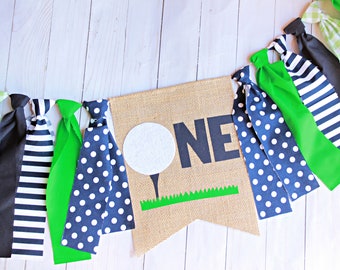 Op de afbeelding: Een banner van jute met het woord "ONE" in zwarte letters en een witte golfbal met een groene steel en gras. De banner is versierd met groene, zwart-wit gestreepte en donkerblauwe banden met witte stippen.
