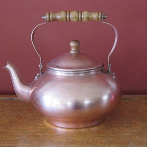 May include: A copper teapot with a wooden handle. The teapot has a shiny, polished finish and a round body.