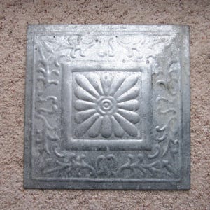 May include: A silver metal ceiling tile with a decorative floral design. The tile has a raised, embossed pattern of a flower with a center circle.