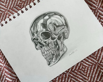 Könnte beinhalten: Eine detaillierte Bleistiftzeichnung eines Schädels mit einem reflektierenden, metallischen Aussehen. Das Kunstwerk befindet sich auf einem weißen, spiralgebundenen Notizblock, mit der Signatur des Künstlers und dem Jahr '25. Der Hintergrund ist ein rot-weiß gemusterter Stoff.