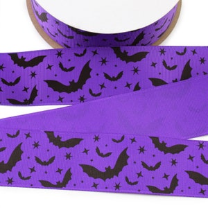 K&ouml;nnte beinhalten: Ein violettes Ripsband mit einem sich wiederholenden Muster aus schwarzen Flederm&auml;usen und kleinen vierzackigen Sternen. Das Band ist auf eine runde Spule gewickelt und weist eine texturierte Oberfl&auml;che mit einem durchgehenden Druck auf.