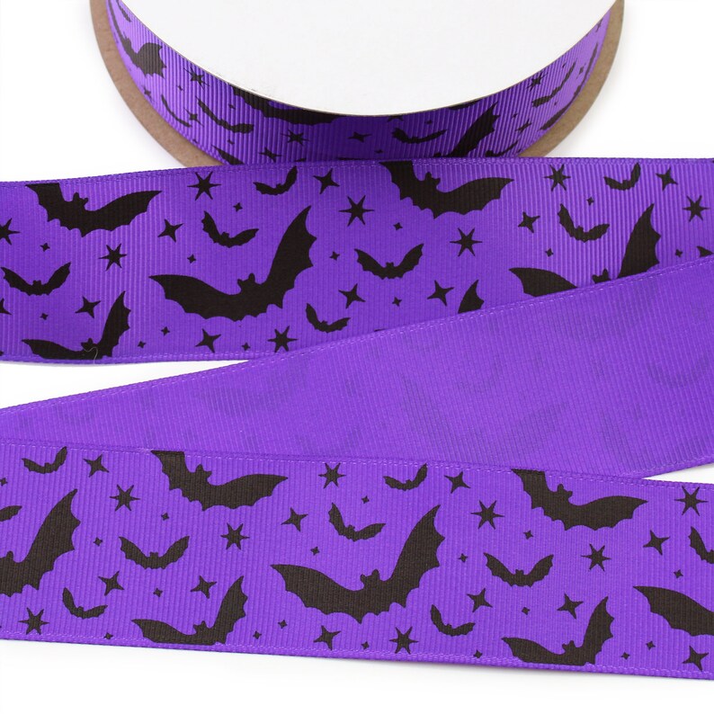 K&ouml;nnte beinhalten: Ein violettes Ripsband mit einem sich wiederholenden Muster aus schwarzen Flederm&auml;usen und kleinen vierzackigen Sternen. Das Band ist auf eine runde Spule gewickelt und weist eine texturierte Oberfl&auml;che mit einem durchgehenden Druck auf.