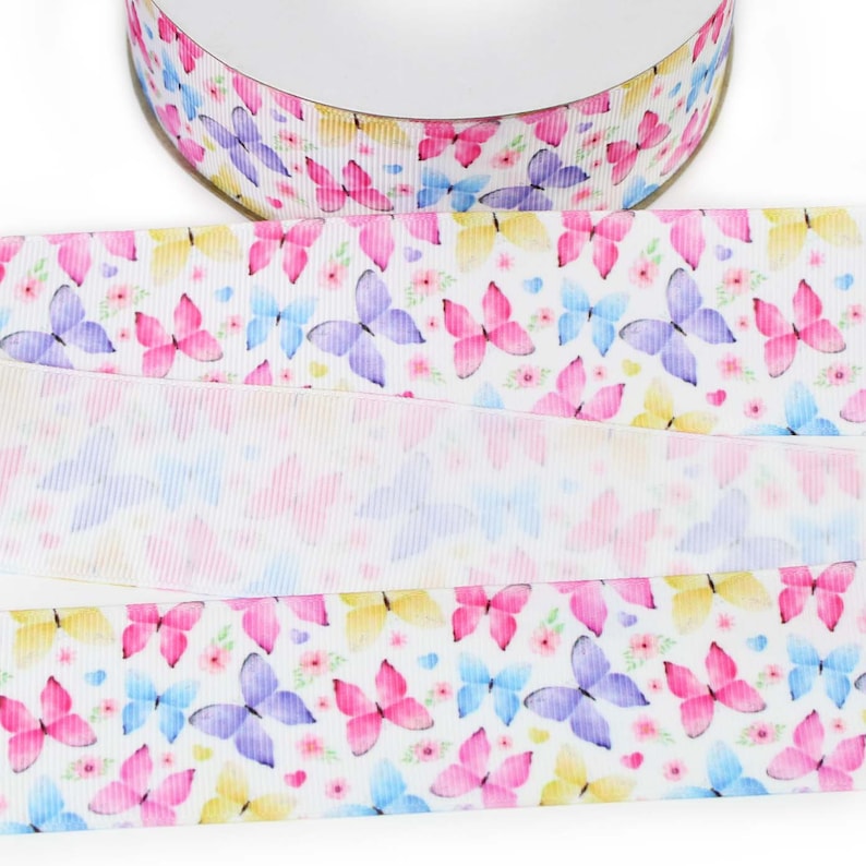 May include: A spool of white grosgrain ribbon featuring a repeating pattern of watercolour-style butterflies in shades of pink, purple, blue, and yellow, interspersed with small pink flowers and tiny blue hearts across the textured fabric surface.