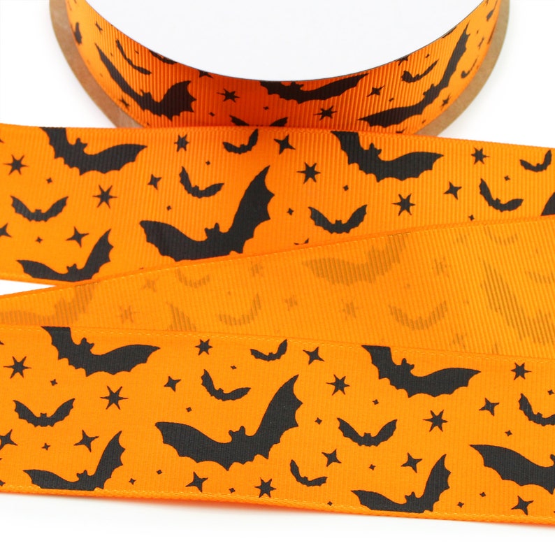 K&ouml;nnte beinhalten: Eine Rolle leuchtend orangefarbenes Ripsband mit einem sich wiederholenden Muster aus schwarzen Silhouetten fliegender Flederm&auml;use und kleinen vierzackigen Sternen. Das Band hat eine ausgepr&auml;gte gerippte Textur und ist auf einen Pappkern gewickelt.