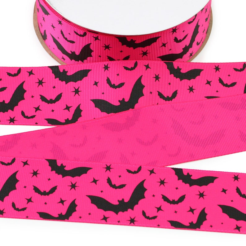 K&ouml;nnte beinhalten: Eine Rolle leuchtend rosa Ripsband mit einem sich wiederholenden Muster aus schwarzen Silhouetten fliegender Flederm&auml;use und kleinen vierzackigen Sternen. Das Band hat eine strukturierte, gerippte Oberfl&auml;che und ist in gefalteter L&auml;nge abgebildet.