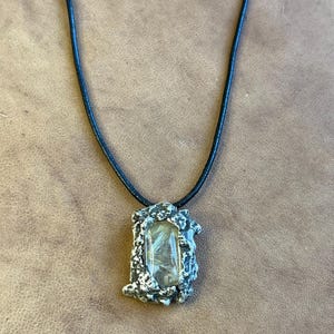 Puede incluir: Un collar con colgante que presenta una piedra rectangular transparente con inclusiones plumosas, engastada en un marco de metal plateado texturizado. Cuelga de un cordón negro. El collar se exhibe sobre una superficie de cuero marrón.