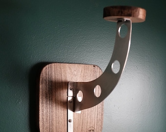 Può includere: Un porta casco a parete con base in legno e braccio in metallo curvo. Il braccio ha tre ritagli circolari e un piccolo ripiano in legno nella parte superiore. Il gancio è in metallo. Il supporto è montato su una parete verde scuro.
