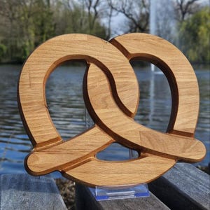 Op de afbeelding: Een houten pretzel-beeldhouwwerk met een natuurlijke houtkleur. Het beeldhouwwerk is in de vorm van een pretzel gebeeldhouwd en wordt getoond op een transparante acrylstandaard.