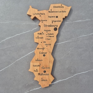 Può includere: Una mappa da parete in legno color marrone chiaro della regione dell'Alsazia in Francia, con nomi di città incisi come Strasburgo, Colmar e Mulhouse, insieme a piccoli punti neri che segnano posizioni specifiche sulla forma geografica.