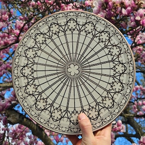 Könnte beinhalten: Eine kreisförmige Holzplatte mit einem detaillierten, schwarz lasergravierten Muster eines gotischen Rosenfensters. Das Design besteht aus strahlenförmigen Speichen, konzentrischen Kreisen und komplexen Vierpassmotiven um eine kleine Blume.