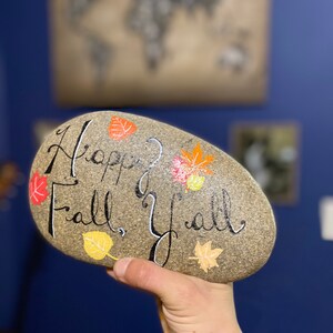 May include: A hand-painted, oval-shaped stone with the words "Happy Fall, Y'all" in black script. Colorful autumn leaves in red, orange, and yellow are painted around the text. The stone is held against a blue background.