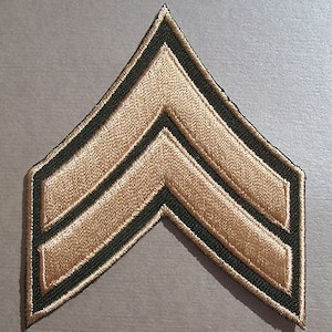 May include: A tan and green military rank patch with three stripes. The patch is made of fabric and has a stitched edge.