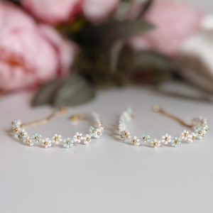 Könnte beinhalten: Zwei zarte Armbänder mit floralem Design. Jedes Armband hat kleine weiße und hellblaue Blüten mit goldfarbenen Akzenten. Die Armbänder liegen auf einer weißen Oberfläche, mit verschwommenen rosa Blüten im Hintergrund.