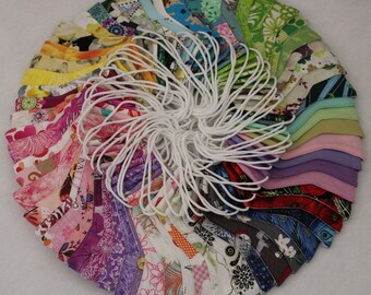 May include: A collection of colorful fabric face masks with various patterns, including floral, geometric, and abstract designs. The masks are arranged in a circle, with the white elastic bands visible.