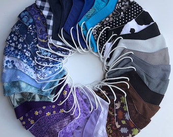 May include: A collection of 20 fabric face masks in various colors and patterns, including blue, purple, gray, black, and white. The masks are arranged in a circle, with the white elastic bands visible.