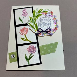 May include: A cream-colored greeting card featuring a vertical black strip containing three floral illustrations: a purple sprig, a pink tulip, and a purple rose. A circular floral wreath with the text "happy birthday" and a purple twine bow sits above.
