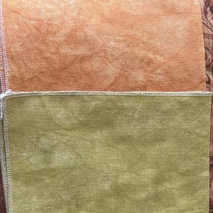 May include: Two pieces of fabric, one above the other. The top piece is a peach color, and the bottom piece is a muted olive green. Both pieces have a woven texture and are edged with white stitching. The fabric appears to be for crafting.