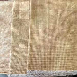 May include: Three rectangular pieces of golden-tan fabric with a visible woven grid texture. Each piece features a mottled, variegated color pattern and is finished with a white serged edge along the bottom, showing a clear, uniform weave.