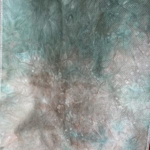 May include: A piece of hand-dyed cross-stitch fabric in shades of teal, brown, and white. The fabric has a textured appearance with a visible grid pattern, and the edges are finished with a white overlock stitch. The overall effect is a watercolor-like blend of colors.