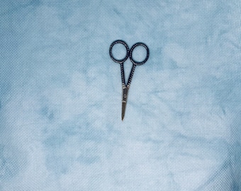 May include: A pair of small, black-handled scissors with silver blades, lying on a light blue fabric background.