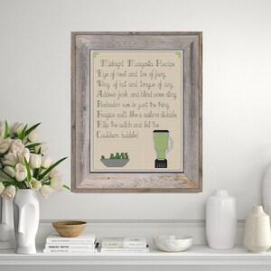 May include: A framed cross-stitch design with a recipe for a "Midnight Margarita" using ingredients like "eye of nest and toe of frog" and "wing of bat and tongue of dog". The design includes a blender and a bowl of green peas.