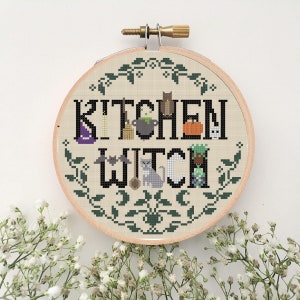 Puede incluir: Un aro de bordado redondo con un fondo de tela beige que presenta un diseño de punto de cruz negro que dice "Kitchen Witch". El diseño incluye varios iconos de temática de Halloween como un caldero, una escoba, un gato y murciélagos. El aro está rodeado por un borde de hojas verdes.