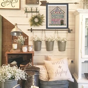 K&ouml;nnte beinhalten: Ein gerahmtes Kreuzstichbild mit einem Hausmotiv und dem Text "In diesem Haus glauben wir: Liebe ist Liebe, Black Lives Matter, Kein Mensch ist illegal, Wissenschaft ist real, Die Erde ist heilig, Frauenrechte sind Menschenrechte, Freundlichkeit ist alles". Der Rahmen h&auml;ngt an einer wei&szlig;en Schiffslappenwand mit einer Metallstange und drei h&auml;ngenden Blument&ouml;pfen.