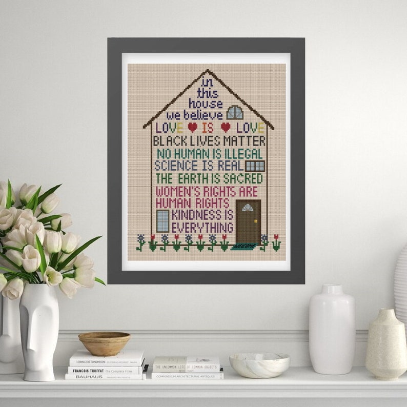 K&ouml;nnte beinhalten: Ein gerahmtes Kreuzstich-Design mit einem Haus, auf dem die Worte "In diesem Haus glauben wir..." und eine Liste von Werten stehen, darunter "Liebe ist Liebe", "Black Lives Matter", "Kein Mensch ist illegal", "Wissenschaft ist real", "Die Erde ist heilig", "Frauenrechte sind Menschenrechte" und "Freundlichkeit ist alles".