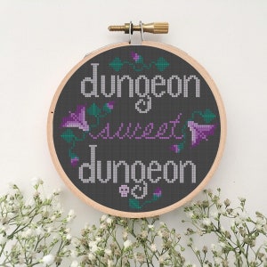 May include: A black embroidery hoop with the text "dungeon & sweet dungeon" in white and purple lettering. The text is surrounded by purple flowers and green leaves.