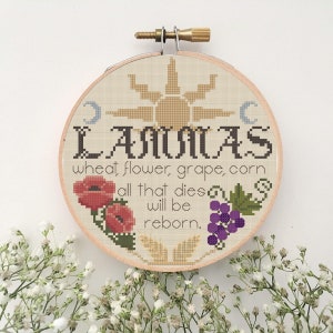 Puede incluir: Un aro de bordado redondo con un fondo beige y un bordado marrón que representa un sol, una luna, trigo, flores, uvas y maíz. El texto "LANMAS wheat flower, grape, corn all that dies will be reborn." está bordado en hilo marrón.