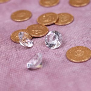 May include: Close-up of clear, faceted gemstones and gold coins on a pink surface. The gemstones are oval-shaped and reflect light, while the coins have a textured surface. The arrangement suggests a theme of luxury or value.