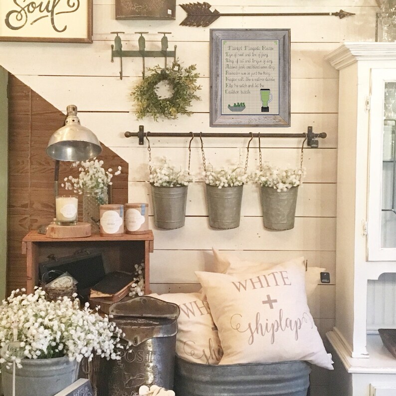 May include: A white farmhouse style wall decor with a framed poem about "Pickle Smoothie Recipe" hanging on the wall.  The poem is written in black text on a white background.  There are three white metal buckets hanging from a metal rod with white flowers in each bucket.  The wall is white shiplap with a brown wooden accent wall.  There is a white pillow with the text "WHITE + shiplap" on it.