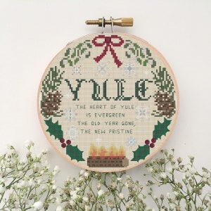 Può includere: Un cerchio da ricamo rotondo in legno con un disegno a punto croce che presenta la parola "YULE" in lettere verdi circondata da una ghirlanda di rami sempreverdi, pigne, agrifoglio e fiocchi di neve. Il testo "THE HEART OF YULE IS EVERGREEN THE OLD YEAR GONE, THE NEW PRISTINE" è ricamato in filo nero sotto la parola "YULE".