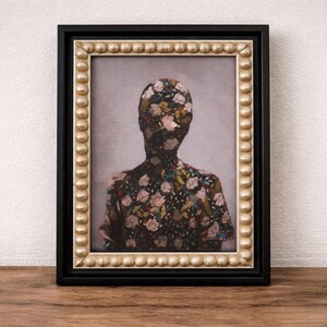 May include: Framed artwork featuring a floral pattern covering the silhouette of a person. The painting is set against a light gray background, framed in black with a gold beaded inner border. The artwork is displayed on a wooden surface.