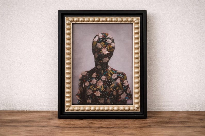 May include: Framed artwork featuring a floral pattern covering the silhouette of a person. The painting is set against a light gray background, framed in black with a gold beaded inner border. The artwork is displayed on a wooden surface.