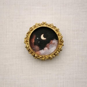 May include: Miniature painting in a gold ornate frame. The artwork depicts a night sky scene with a crescent moon, stars, and clouds in shades of black, brown, and white. The frame has a detailed, vintage-style design.