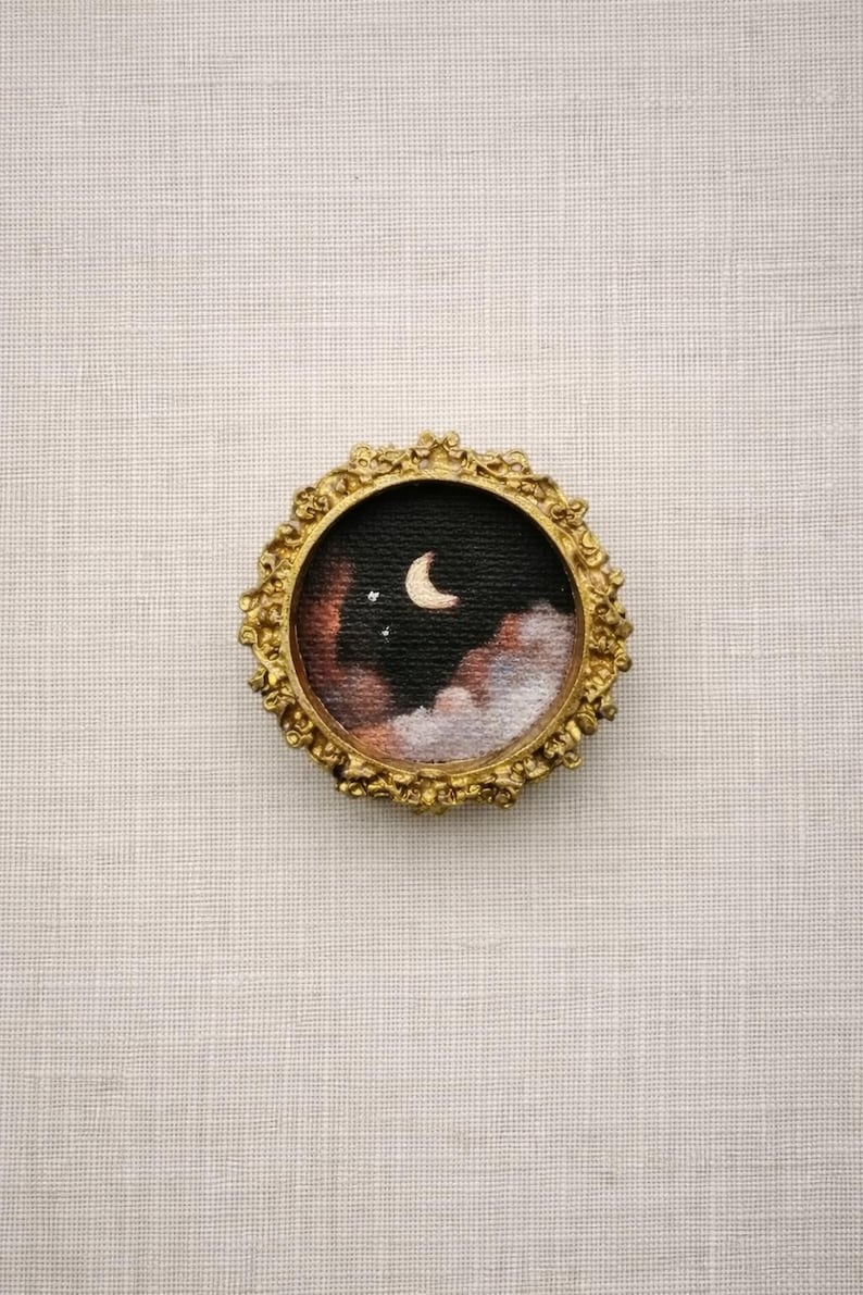 May include: Miniature painting in a gold ornate frame. The artwork depicts a night sky scene with a crescent moon, stars, and clouds in shades of black, brown, and white. The frame has a detailed, vintage-style design.